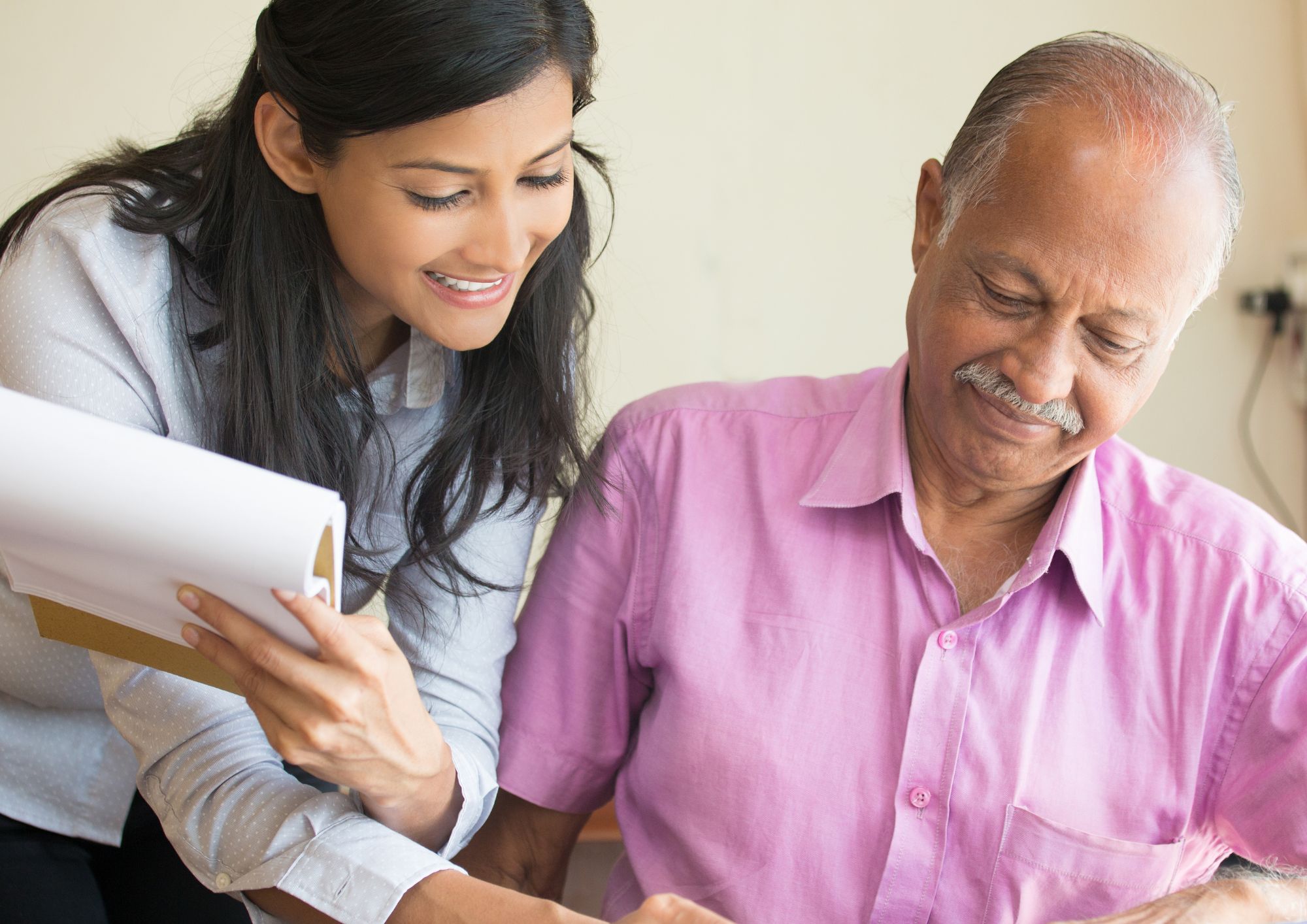 Woman holding a pad reads something that an older man, who could be her father, is reading. they are smiling.