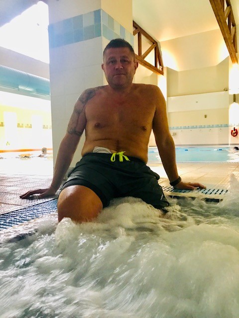 A man is sat with his legs in the pool. A visible stoma pouch is seen at the top of the waist band of hi swimming shorts.