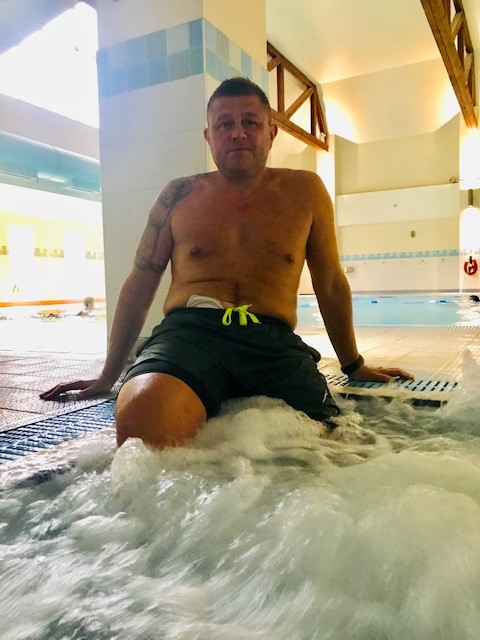 A man is sat with his legs in the pool.  A visible stoma pouch is seen at the top of the waist band of hi swimming shorts.