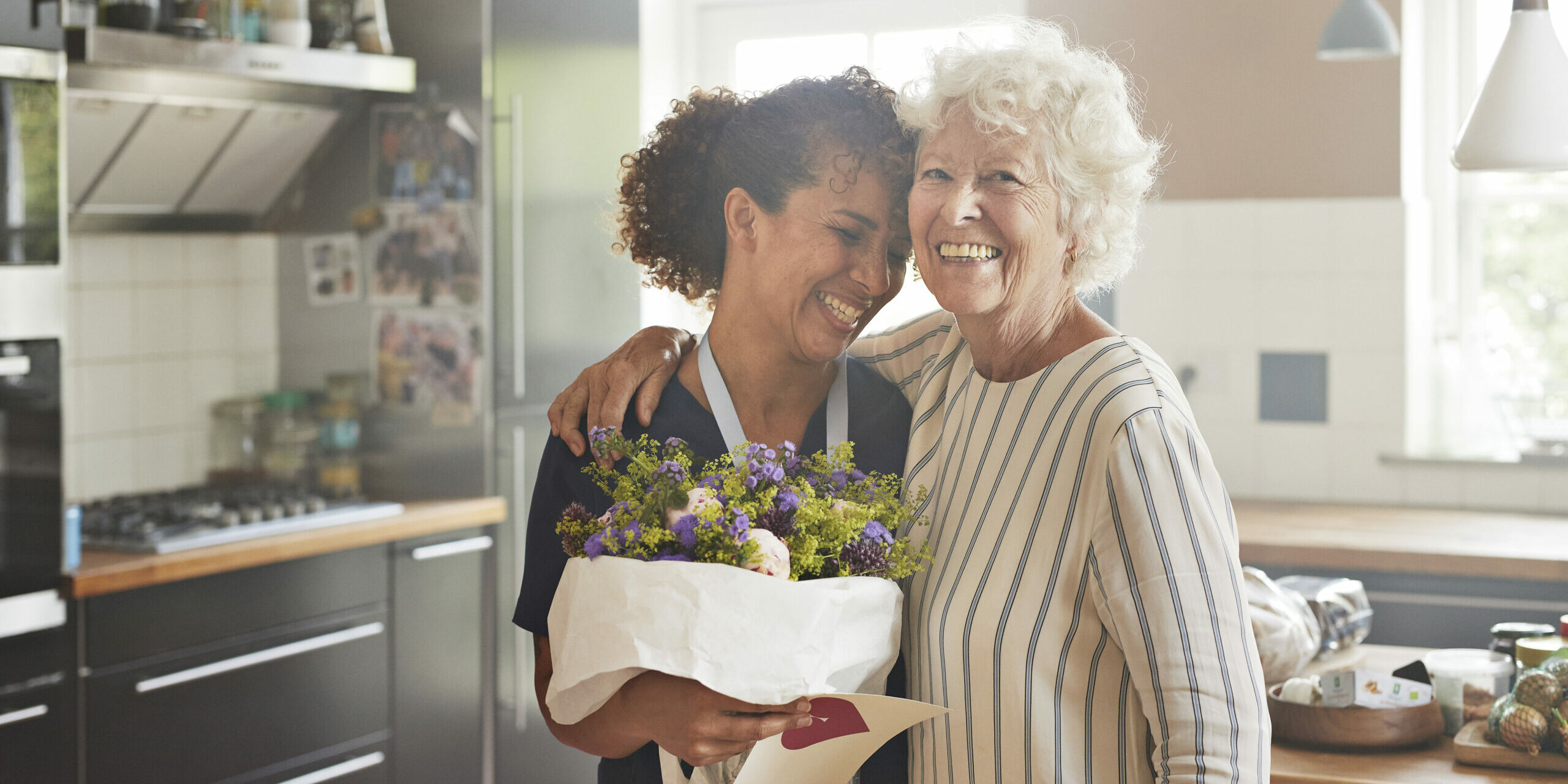 Happy senior woman with arm around female caregiver in kitchen