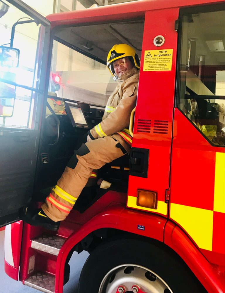 A man is sat in his firefighter uniform and is sat in a seat of a fire engine looking at the camera and smiling