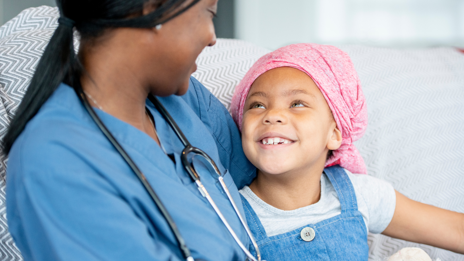 A nurse and a child sat next to each other. The child is looking at the nurse and grinning. The image is to illustrate why bedwetting should be treated.