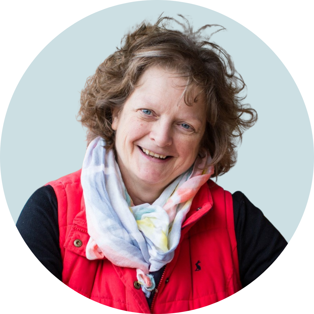 Smiling woman with short curly brown hair wearing a red gilet and multicoloured scarf over a black top, in front of a pale blue background.