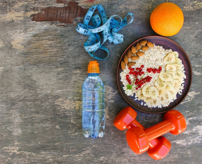 A healthy lifestyle flat lay featuring a bowl of oats with banana, almonds, and redcurrants, a water bottle, orange dumbbells, an orange, and a blue measuring tape on a wooden surface.