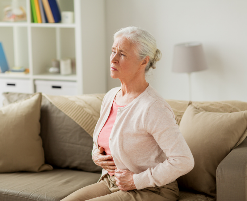 An older woman sitting on a sofa, holding her stomach with a pained expression, suggesting discomfort or abdominal pain.