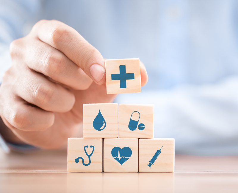 Hand stacking wooden blocks featuring healthcare-related icons including a stethoscope, heart monitor, syringe, pill, water droplet, and a medical cross.