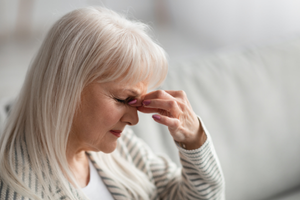 Elderly woman with long white hair pressing her fingers to the bridge of her nose, showing discomfort, seated indoors.