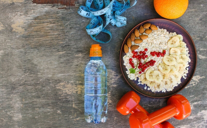 A healthy lifestyle flat lay featuring a bowl of oats with banana, almonds, and redcurrants, a water bottle, orange dumbbells, an orange, and a blue measuring tape on a wooden surface.