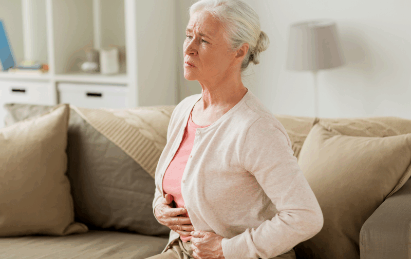 An older woman sitting on a sofa, holding her stomach with a pained expression, suggesting discomfort or abdominal pain.