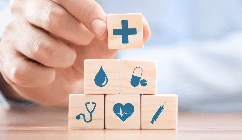Hand stacking wooden blocks featuring healthcare-related icons including a stethoscope, heart monitor, syringe, pill, water droplet, and a medical cross.
