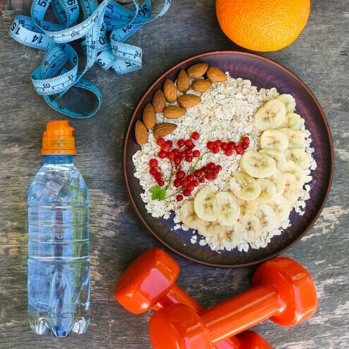 A healthy lifestyle flat lay featuring a bowl of oats with banana, almonds, and redcurrants, a water bottle, orange dumbbells, an orange, and a blue measuring tape on a wooden surface.