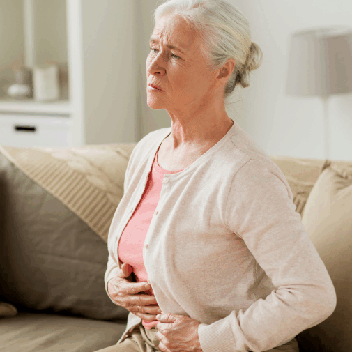 An older woman sitting on a sofa, holding her stomach with a pained expression, suggesting discomfort or abdominal pain.