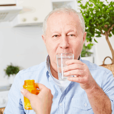 An older man sitting in a kitchen, holding a glass of water while someone hands him a pill bottle, implying support with taking medication.