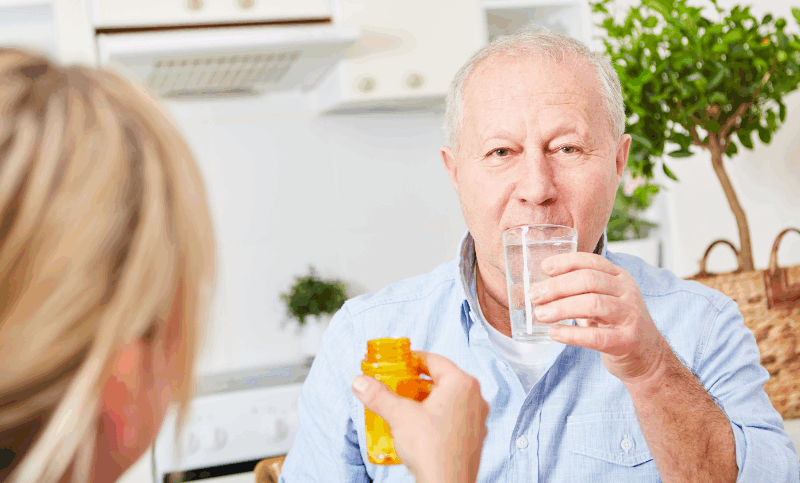 An older man sitting in a kitchen, holding a glass of water while someone hands him a pill bottle, implying support with taking medication.