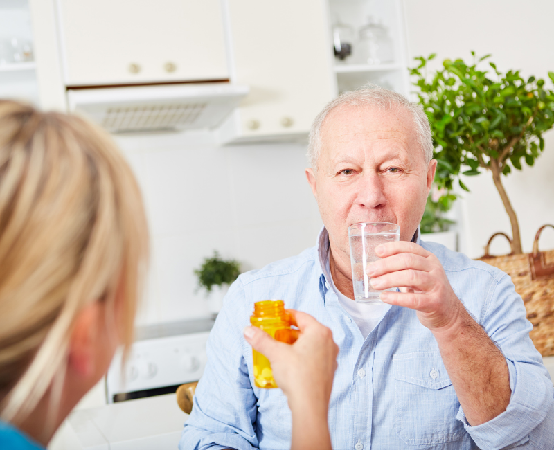 An older man sitting in a kitchen, holding a glass of water while someone hands him a pill bottle, implying support with taking medication.