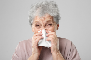 Older woman with short grey hair wearing a pale pink top, sneezing into a tissue against a neutral grey background.