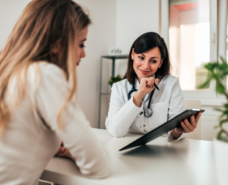 A female doctor smiling and holding a clipboard while speaking with a female patient across the desk in a bright consultation room.