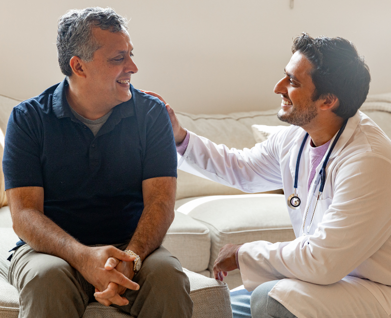 Smiling middle-aged man sitting on a sofa receiving friendly reassurance from a male doctor in a white coat with a stethoscope, both making eye contact and laughing warmly.