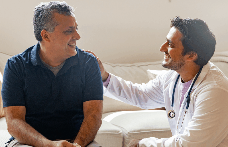 Smiling middle-aged man sitting on a sofa receiving friendly reassurance from a male doctor in a white coat with a stethoscope, both making eye contact and laughing warmly.