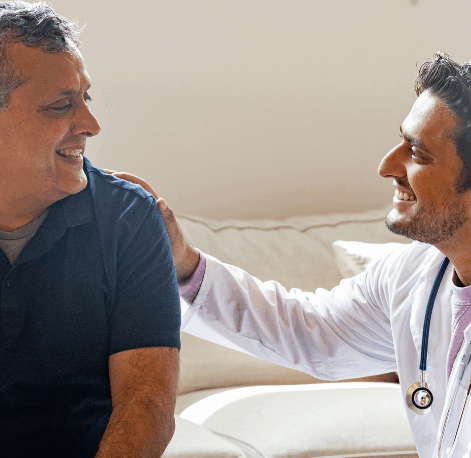 Smiling middle-aged man sitting on a sofa receiving friendly reassurance from a male doctor in a white coat with a stethoscope, both making eye contact and laughing warmly.