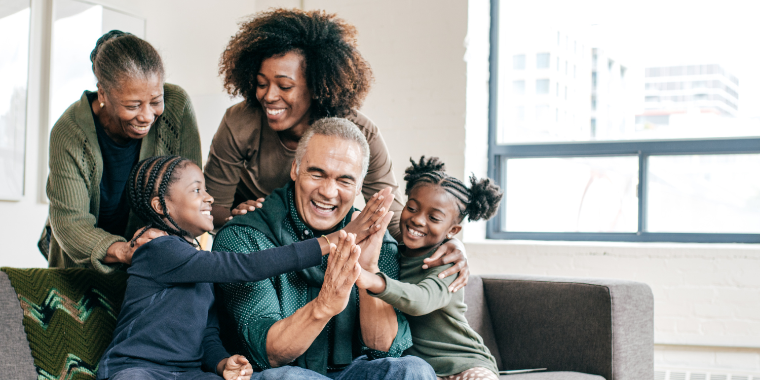 “A joyful multi-generational family sitting together on a sofa, smiling and laughing as two young girls high-five an older man.